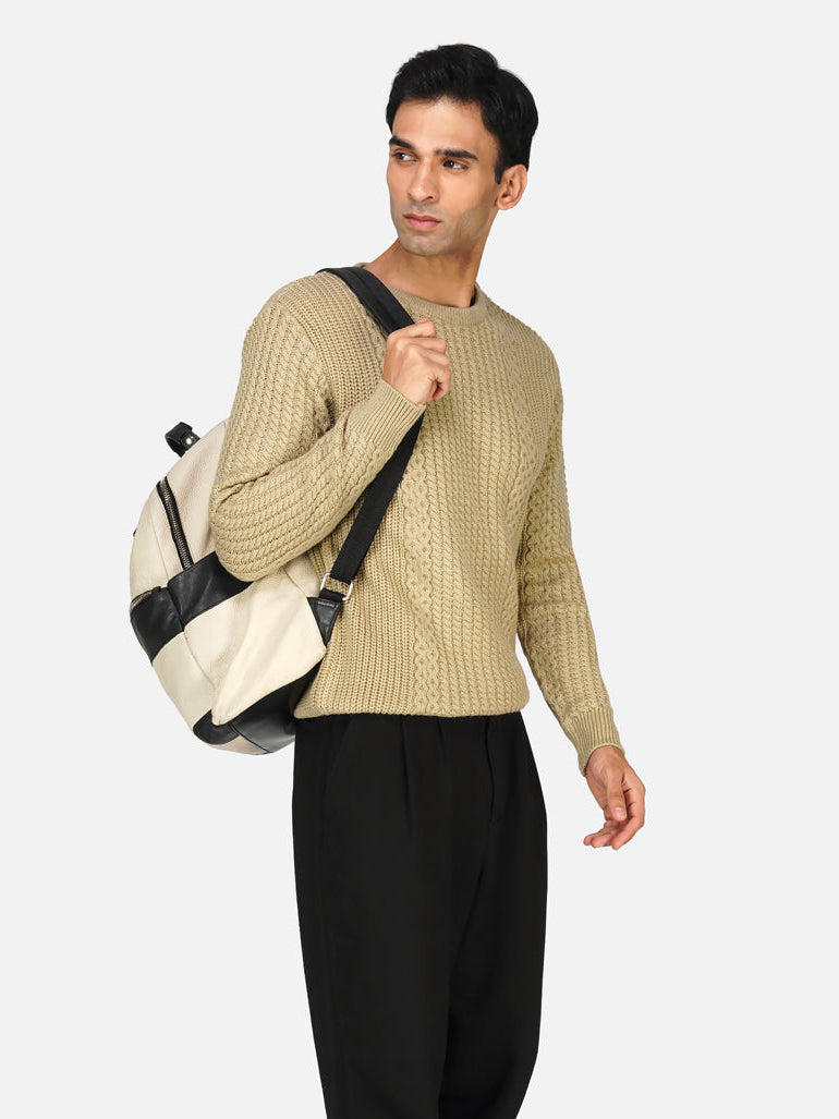 A male model wearing a beige cable-knit sweater and black trousers, carrying a white bag.